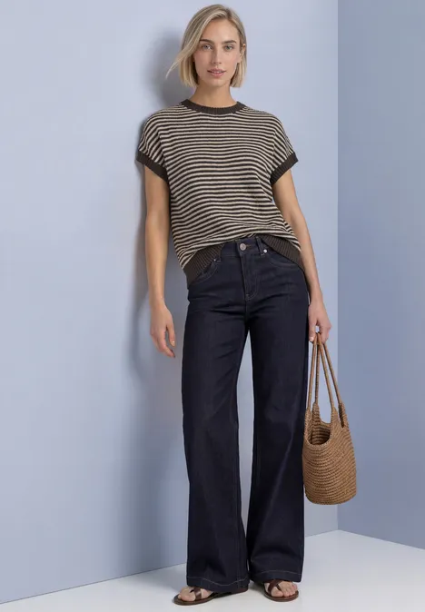 Frau in gestreiftem T-Shirt, weiten Jeans und Strohtasche vor hellgrauer Wand.