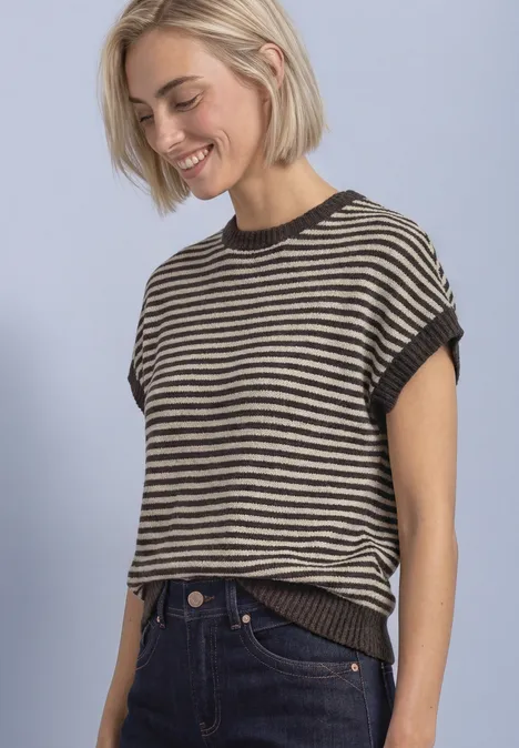 Eine blonde Frau lächelt in einem gestreiften Kurzarmshirt und Jeans vor hellem Hintergrund.