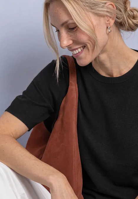 Junge Frau mit blondem Haar trägt schwarzes T-Shirt und braune Schultertasche, lächelt seitlich.