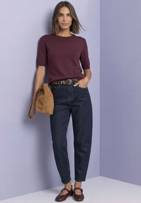Femme en pull violet à manches courtes, jean foncé, ballerines marron et sac beige devant un mur lilas.
