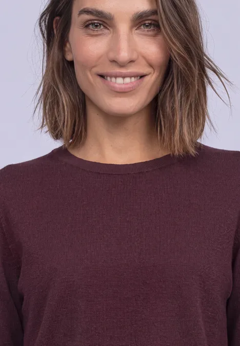 Gros plan d’une femme souriante aux cheveux mi-longs portant un haut bordeaux sur fond clair