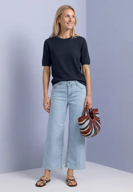 Frau in schwarzem T-Shirt, weiten, hellen Jeans und Sandalen hält gestreifte Handtasche.