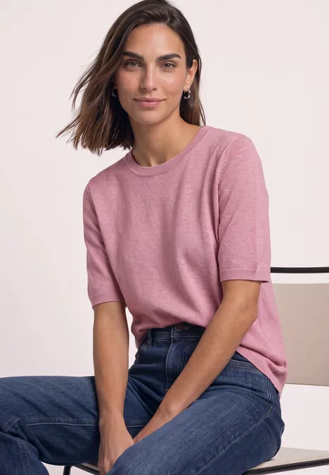 Vrouw in roze shirt en jeans zit en glimlacht.