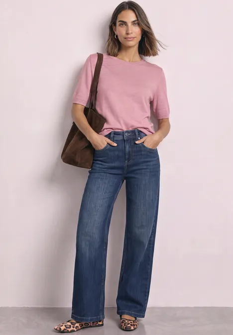Vrouw in roze T-shirt en wijdvallende jeans, met een bruine tas, staand voor een lichte achtergrond.