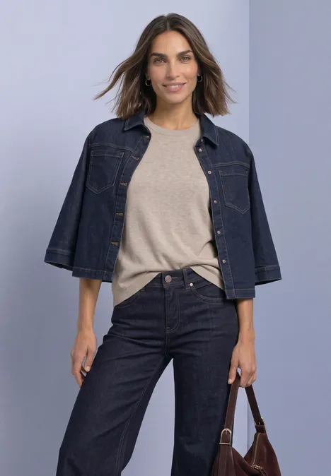 Eine lächelnde Frau trägt helle Bluse, dunkelblaue Jeansjacke mit Jeanshose und hält eine braune Tasche.