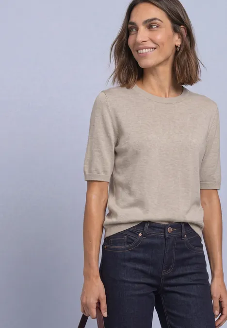 Frau trägt beige Oberteil mit Ellbogenärmeln und dunkle Jeans vor schlichtem Hintergrund.