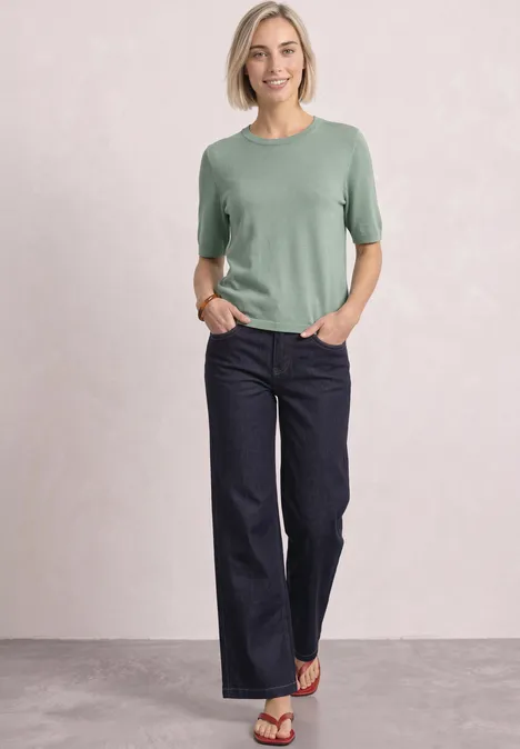 Kurzarm Pullover mit Rundhals in Unifarbe eucalyptus green Frau in grünem T-Shirt und weitem dunklem Jeans, steht entspannt mit Händen in den Taschen vor hellem Hintergrund.