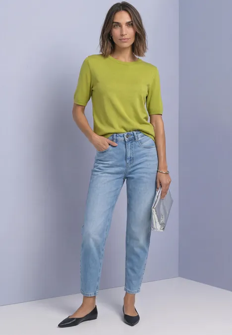 Frau in grünem Kurzarmshirt und blauen Jeans, hält silberne Clutch und trägt schwarze Ballerinas.