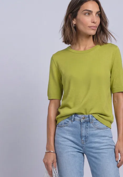 Eine Frau mit schulterlangem braunem Haar trägt ein hellgrünes T-Shirt und blaue Jeans vor hellem Hintergrund.