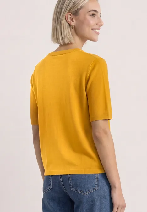 Frau von hinten in senfgelbem Kurzarmshirt und blauen Jeans, lächelnd seitlich blickend.