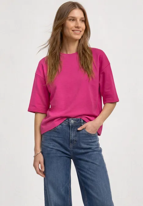 Uśmiechnięta kobieta w różowym t-shircie i niebieskich dżinsach, swobodna poza.