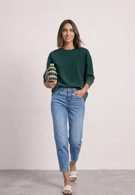 Kurzarm Sweatshirt mit Rundhals shadow teal Frau in grünem Pullover, blauen Jeans und weißen Sandalen steht vor neutralem Hintergrund und lächelt.
