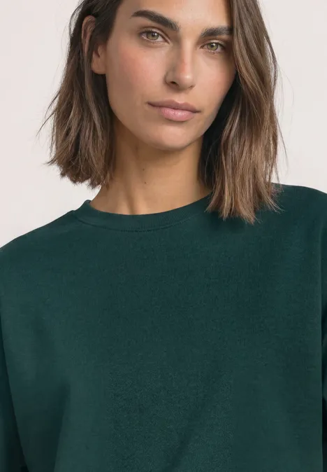 Kurzarm Sweatshirt mit Rundhals shadow teal Porträt einer Frau mit schulterlangen Haaren und dunkelgrünem Pullover vor neutralem Hintergrund.