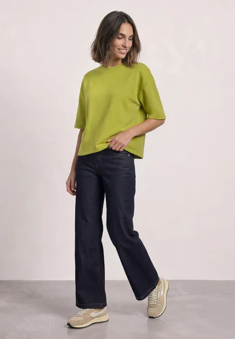 Kurzarm Sweatshirt mit Rundhals apple lime Eine Frau in einer grünen Bluse, weiten Jeans und Plateau-Sneakern posiert vor hellem Hintergrund.