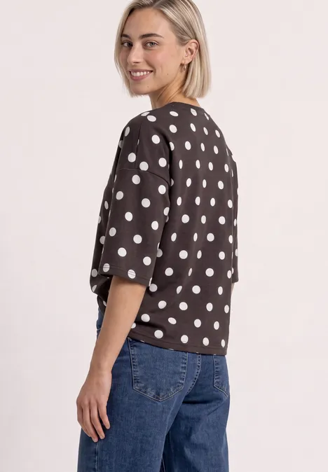 Sweatshirt met korte mouwen en ronde hals black coffee Frau im braunen, weiß gepunkteten Oberteil mit kurzen Ärmeln und blauen Jeans, steht halb gewandt lächelnd.