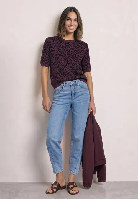 Frau in hoher Taillenjeans, dunkelrotem Shirt und Sandalen, hält Jacke in der Hand.