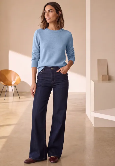 Vrouw in minimalistische ruimte draagt lichtblauwe trui, donkere wijd uitlopende jeans en bruine schoenen.