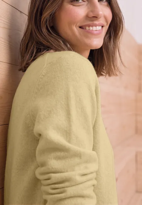 Trui met lange mouwen en boothals sunbeam yellow mel. Eine lächelnde Frau in hellem Pullover lehnt sich entspannt an eine Holzvertäfelung.