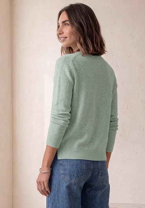 Trui met lange mouwen en boothals eucalyptus green mel. Eine Frau in grünem Pullover und Jeans steht seitlich, Blick über die Schulter, vor hellem Hintergrund.
