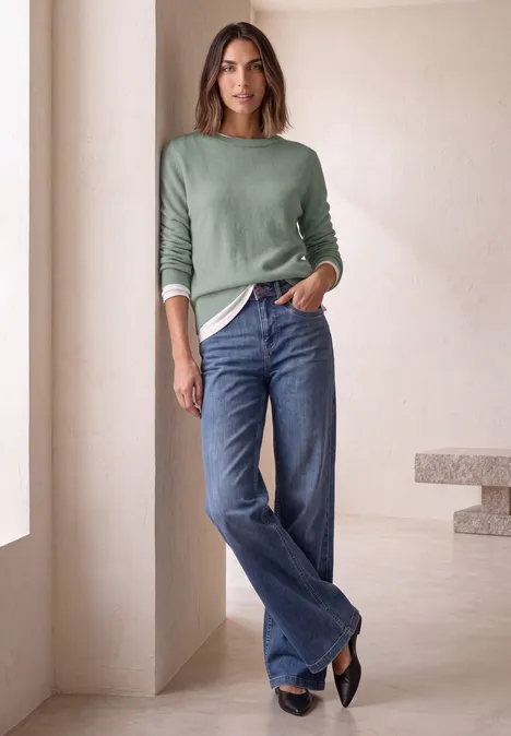 Trui met lange mouwen en boothals eucalyptus green mel. Frau in grünem Pullover und weitem Jeansmodell lehnt lässig in hellem, minimalistischem Raum.