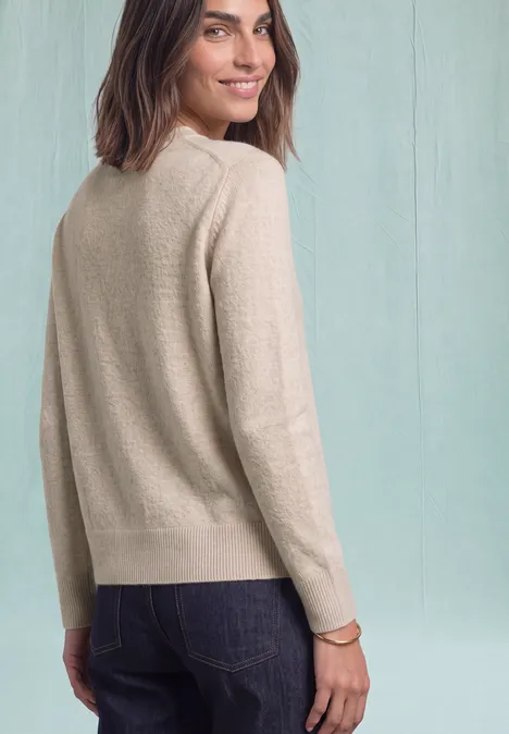 Vrouw in beige trui en spijkerbroek kijkt over haar schouder en glimlacht licht.