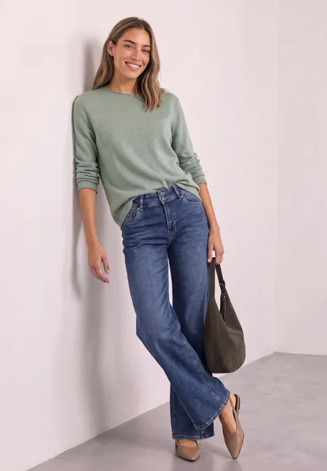 Pullover mit Boat Neck und Rollkanten eucalyptus green mel. Junge Frau lehnt lächelnd an weißer Wand, trägt grünen Pullover, blaue Jeans und hält eine Tasche.
