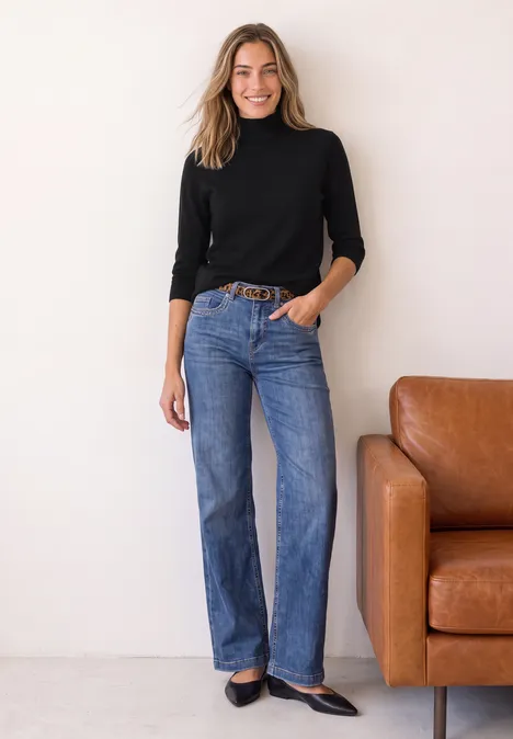 Pull basique à col roulé Black Eine Frau in schwarzem Rollkragenpullover und blauen Jeans steht lässig neben einem braunen Ledersofa.