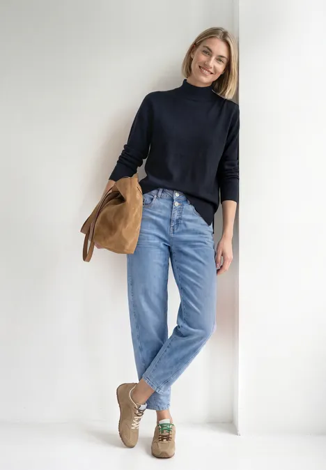 Basic sweter z golfem darkest blue Uśmiechnięta kobieta oparta o biały mur, ma czarny golf, niebieskie dżinsy i trzyma beżową zamszową kurtkę.