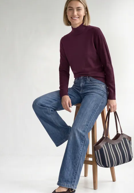 Basic sweter z golfem berry maroon Kobieta z krótkimi blond włosami w fioletowym golfie i szerokich dżinsach siedzi na stołku, trzyma pasiastą torebkę