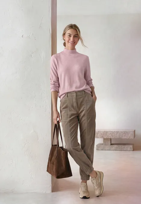 Basic sweter z golfem quiet rose mel. Kobieta w pastelowo różowym swetrze, beżowych spodniach, jasnych botkach i brązowej torbie na prostym tle.