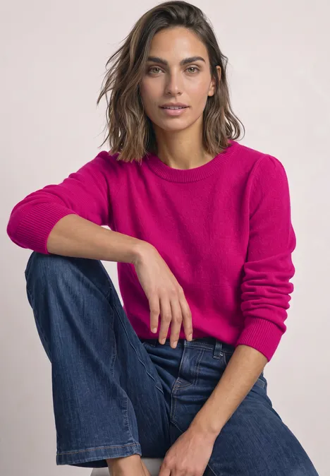 Femme en pull rose et jean, assise jambes croisées, souriante, regardant l'appareil photo.