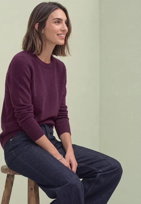 Langarm Pullover mit Rundhals und Raffung berry maroon mel. Eine lächelnde Frau mit schulterlangen braunen Haaren sitzt entspannt auf einem Hocker, trägt Burgunderpullover und Jeans.