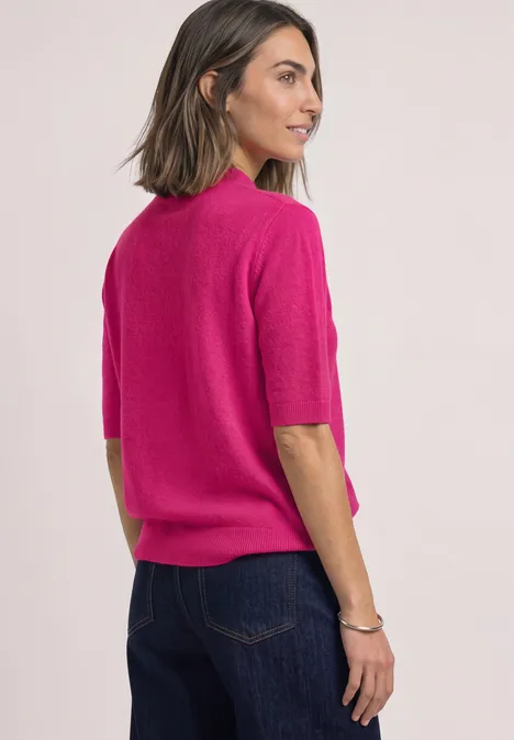 Vrouw met halflang haar in fuchsia kort shirt en blauwe jeans, kijkt schuin opzij.