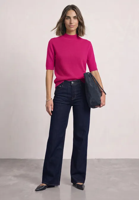 Een vrouw draagt een fuchsia top, wijde jeans en zwarte schoenen en houdt een clutch vast.