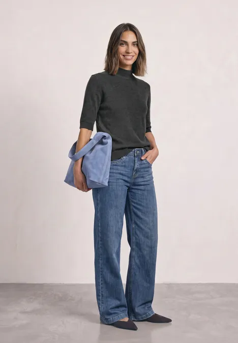 Kurzarm Pullover mit Turtle Neck armor grey mel. Frau in weiten Jeans und schwarzem Pulli, hellblaue Jacke lässig über der Schulter, lächelnd.