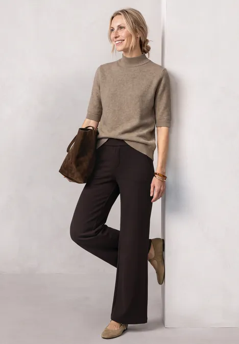 Kurzarm Pullover mit Turtle Neck sanded amber mel. Frau mit beigem Pullover, schwarzen Hosen und grünen Stiefeletten lehnt lächelnd an weißer Wand.