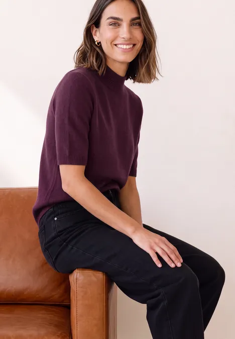 Kurzarm Pullover mit Turtle Neck berry maroon mel. Eine lächelnde Frau sitzt auf der Sofalehne, trägt ein kurzärmeliges violettes Oberteil und schwarze Hose.