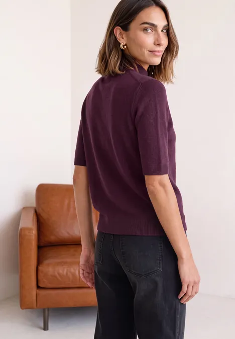 Kurzarm Pullover mit Turtle Neck berry maroon mel. Frau im purpurfarbenen Kurzarm-Pullover und Jeans steht vor weißem Hintergrund neben einem beigen Ledersessel.