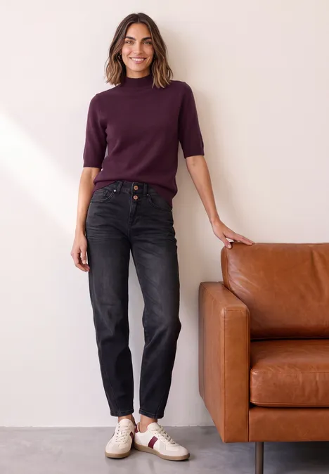 Kurzarm Pullover mit Turtle Neck berry maroon mel. Frau lehnt an braunem Ledersofa, trägt lila Pullover, schwarze Jeans und weiße Sneaker und lächelt.