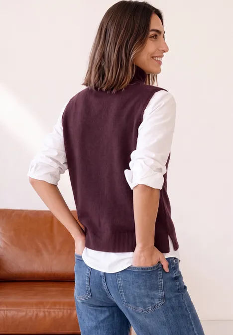 Vrouw draagt witte blouse, paarse gilet en spijkerbroek, kijkt glimlachend opzij.