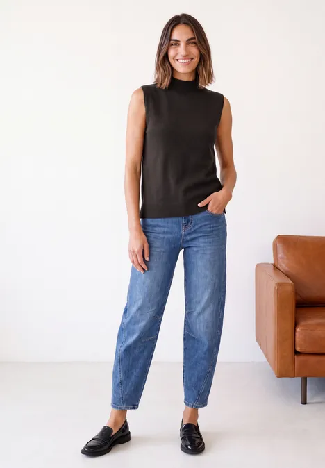 Een glimlachende vrouw in een zwart mouwloos topje, blauwe jeans en zwarte loafers staat voor een bruine leren bank.