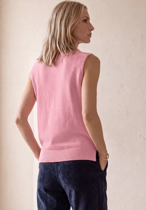 Slanke vrouw in profiel, draagt een roze mouwloos topje en donkere jeans, staand voor een lichte achtergrond.