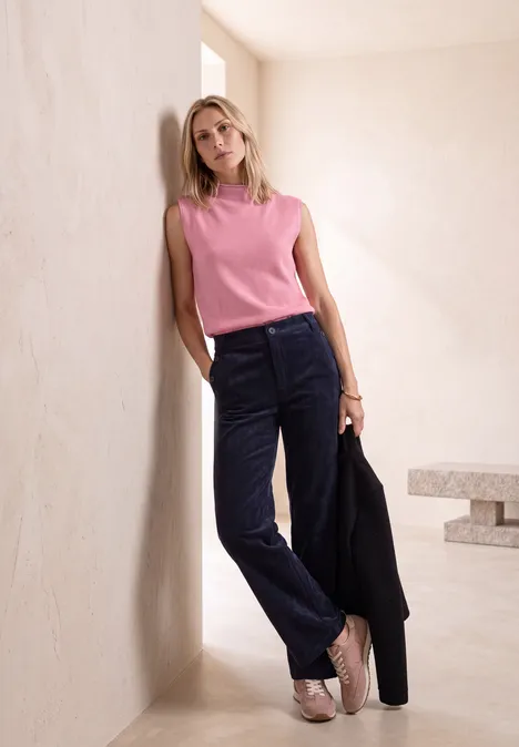 Blonde vrouw leunt tegen een beige muur, roze top, donkerblauwe corduroy broek, zwarte blazer in de hand.