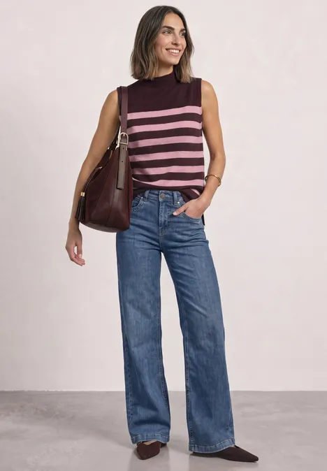 Frau trägt ärmelloses, gestreiftes Top und weite Jeans, steht lächelnd mit Schultertasche.