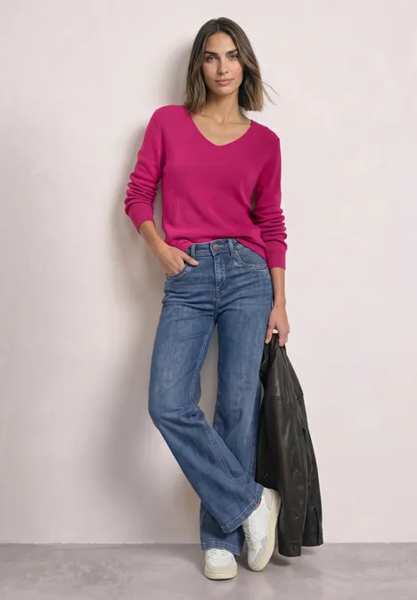 Langarm Pullover mit V-Ausschnitt magenta dream Frau in pinkem Pullover, weiten Jeans und weißen Sneakern hält schwarze Lederjacke vor hellem Hintergrund.