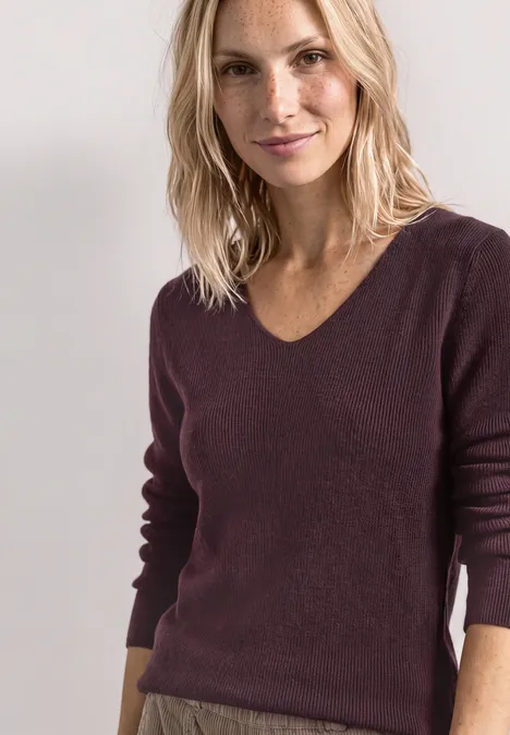 Femme blonde en pull bordeaux devant un fond clair, souriante et posant de façon décontractée