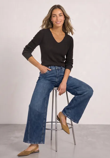 Langarm Pullover mit V-Ausschnitt black coffee Eine Frau sitzt lächelnd auf einem Hocker, trägt schwarzen V-Pullover, weite Jeans und beige flache Schuhe.