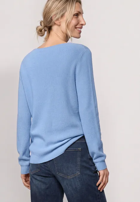 Langarm Pullover mit V-Ausschnitt northern blue Frau steht mit dem Rücken zur Kamera, trägt hellblauen Pullover und Jeans, lächelt seitlich.