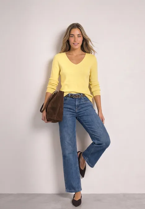Langarm Pullover mit V-Ausschnitt sunbeam yellow Frau in gelbem Pullover, blauen Jeans und brauner Tasche lächelt vor neutralem Hintergrund.