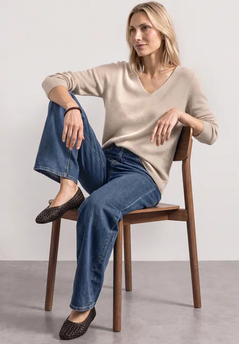 Femme assise sur une chaise, pull beige, jean bleu et ballerines marron, posture détendue.